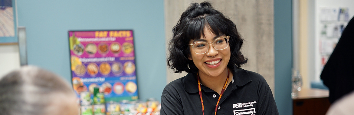 college student with black hair, glasses, and a black ASU shirt sitting in a classroom by a colorful poster