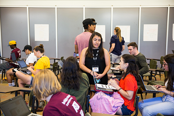 college students talking, working on laptops, and sitting at desks in a classroom
