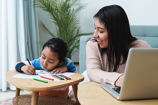 dark-haired woman working at a laptop, looking at a dark-haired girl drawing on a piece of paper