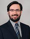 David Androff headshot - fair-skinned man, smiling, black hair and beard, glasses, gray collared shirt, black blazer, blue striped tie