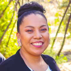 Tahnee Baker headshot - medium-skinned woman, smiling, black hair, black blazer, earrings